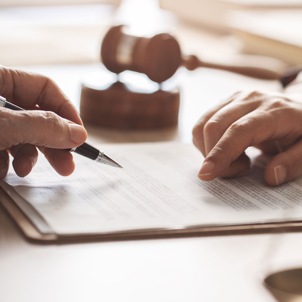 Gros plan sur des mains pointant et signant un document juridique sur un bureau, avec un marteau de juge flou à l’arrière-plan, illustrant la rédaction et la signification d’actes.